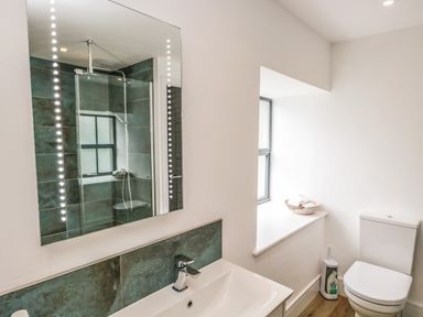A bathroom with a sink and faucet a mirror shower area a toilet and a window with a basket of toilet paper at Bank Hurst Coach House in Ulverston