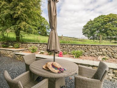 An outdoor seating area with a round wicker table and four wicker chairs with a closed umbrella and straw hats on the table at Bank Hurst Coach House in Ulverston