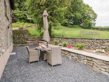 An outdoor patio area with a round table, four wicker chairs, a closed umbrella, and a stone wall with plants at Bank Hurst Coach House in Ulverston