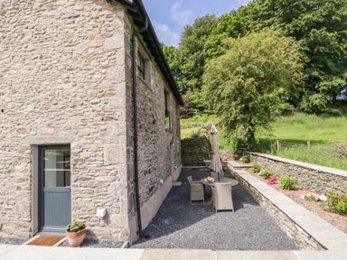 Stone exterior wall of a house with a door opening to a patio area with table chairs umbrella and garden beds at Bank Hurst Coach House in Ulverston