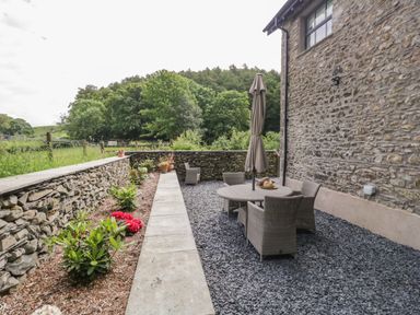 An outdoor patio area with a round table and four chairs under a closed umbrella beside a stone wall and garden beds at Bank Hurst Coach House in Ulverston