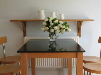 A dining area with a black topped wooden table surrounded by wooden chairs and a shelf with three white pitchers on the wall at House on the Green in Chipping Norton