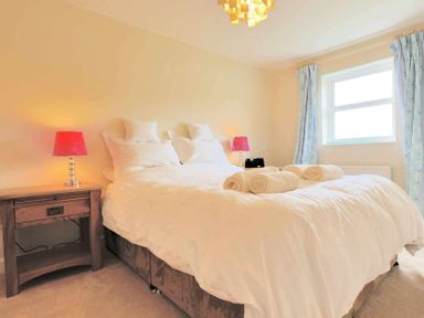 A bedroom with a large bed white bedding and rolled towels on it two bedside tables with red lamps and a window with curtains at House on the Green in Chipping Norton
