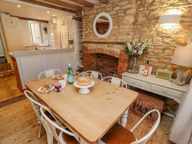 A dining area with a wooden table set with food and drinks beside a stone wall with a fireplace and a side table at Chippy Cottage in Chipping Norton