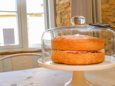 A sponge cake with cream and jam on a white cake stand under a glass dome in a room with stone walls at Chippy Cottage in Chipping Norton