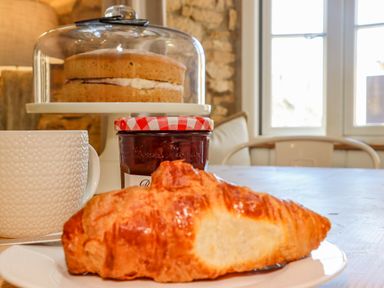 A croissant on a plate with a white mug jam jar and a cake under a glass dome on a wooden table