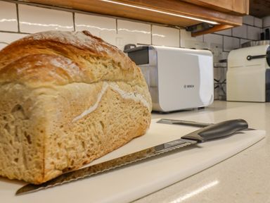 A loaf of bread with a bread knife on a cutting board in a kitchen with a toaster and appliances at Chippy Cottage in Chipping Norton
