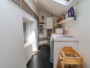 A narrow utility room with a washing machine, microwave, iron, and wooden high chair at Chippy Cottage in Chipping Norton