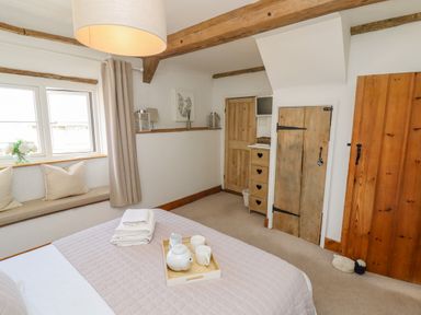 A bedroom with a bed tray holding a teapot and cups a window seat with cushions wooden doors and a small chest of drawers at Chippy Cottage in Chipping Norton
