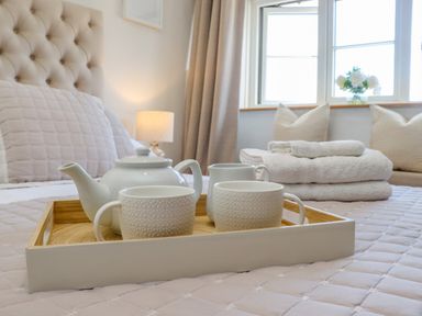 A bedroom with a tufted headboard a tray with a teapot and cups on the bed and folded towels near a window at Chippy Cottage in Chipping Norton