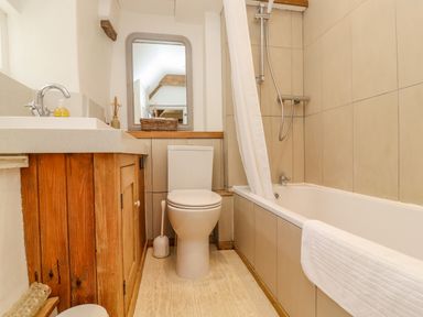 A bathroom with a wooden cabinet, sink, toilet, bathtub with shower, and a mirror above the toilet at Chippy Cottage in Chipping Norton