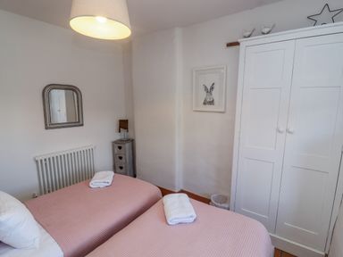 A bedroom with two twin beds with pink covers a white wardrobe a small bedside table and a wall mirror at Chippy Cottage in Chipping Norton
