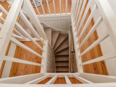 A view looking down a narrow carpeted staircase with white wooden railings and wooden floor at the top at Chippy Cottage in Chipping Norton