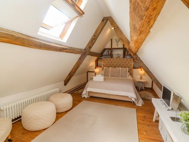 A bedroom with sloped ceiling exposed wooden beams a bed two ottomans a rug and a TV at Chippy Cottage in Chipping Norton