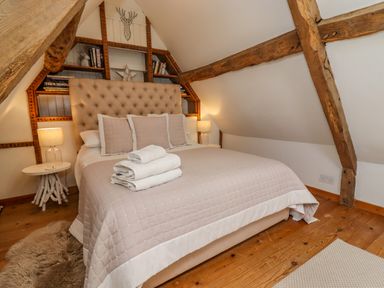 A bedroom with a bed with beige bedding and pillows two bedside tables with lamps wooden beams and built in shelves with books at Chippy Cottage in Chipping Norton