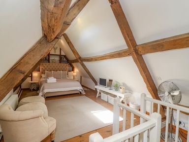 A bedroom with exposed wooden beams a bed with bedside tables and lamps a white bench with a television and fan at Chippy Cottage in Chipping Norton