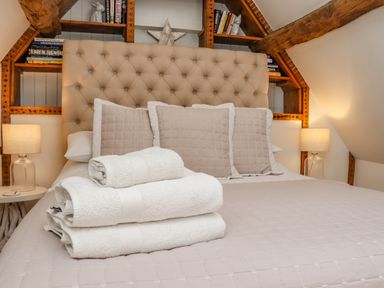 A bedroom with a beige upholstered headboard beige pillows a folded stack of white towels on the bed and wooden shelves with books and decorations at Chippy Cottage in Chipping Norton