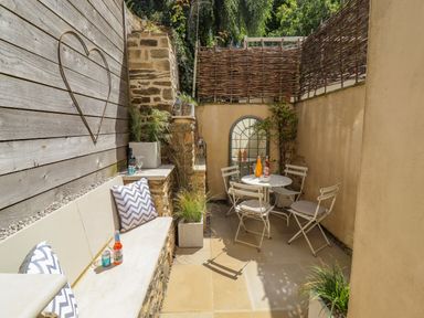 A small outdoor patio with a stone bench with cushions a round table with four chairs and plants at Chippy Cottage in Chipping Norton