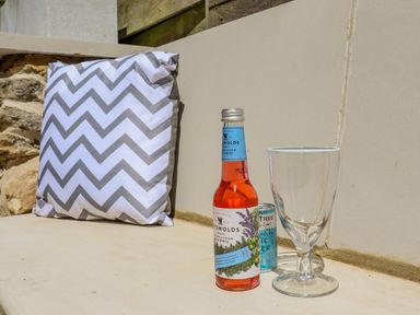 A cushion with a chevron pattern a bottle of Cotswolds drink a can of Fever Tree tonic water and an empty glass on a stone bench at Chippy Cottage in Chipping Norton