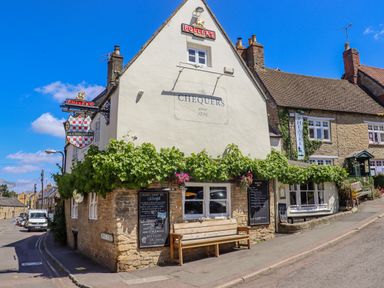 Exterior of a pub named The Chequers with a bench outside and a sign pointing to a theatre in Chipping Norton
