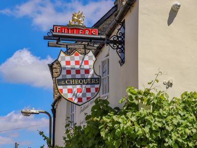 A pub sign for The Chequers with a blue sky background and greenery on the wall at Chippy Cottage in Chipping Norton