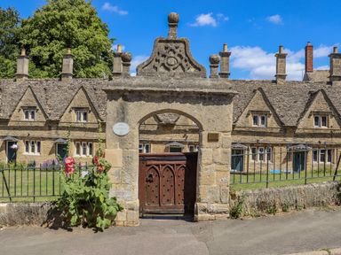 Stone archway with a wooden gate in front of a row of stone cottages with multiple chimneys and a garden with flowers