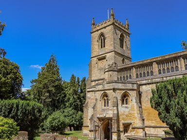 An old stone church with a tall tower and arched windows surrounded by trees and gravestones at Chippy Cottage in Chipping Norton