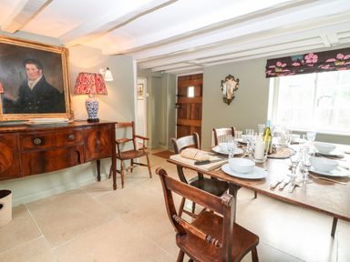 A dining room with a wooden table set with plates glasses and a loaf of bread a wooden sideboard with a lamp and a portrait on the wall at Holly Cottage in Minster Lovell