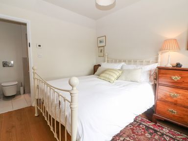 A bedroom with a white metal bed frame green pillows a wooden chest of drawers with a lamp and an adjacent bathroom at Holly Cottage in Minster Lovell