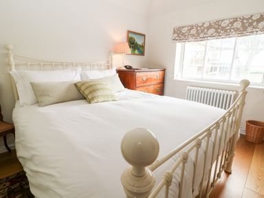 A bedroom with a white metal bed frame a wooden chest of drawers and a window with floral curtains at Holly Cottage in Minster Lovell