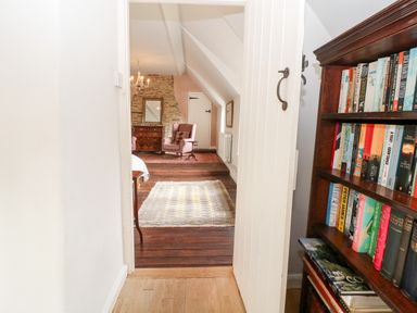 A hallway leading into a living room with wooden floors bookshelves and a pink armchair at Holly Cottage in Minster Lovell
