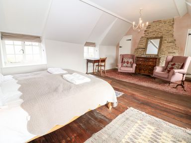 A bedroom with a bed a wooden desk two armchairs a chest of drawers and exposed stone wall at Holly Cottage in Minster Lovell