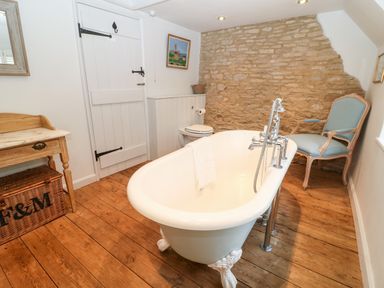 A bathroom with a clawfoot bathtub a wooden floor a blue upholstered chair a wicker basket and a stone wall at Holly Cottage in Minster Lovell