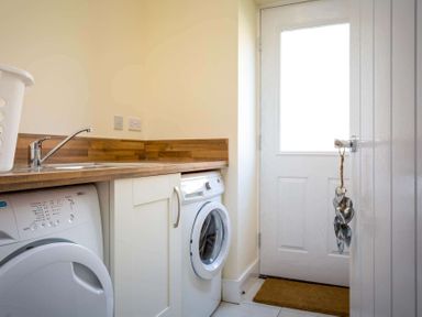 A utility room with a washing machine dryer sink with wooden countertop and a door with a heart shaped hanging at The Bybrook in Bourton-on-the-Water
