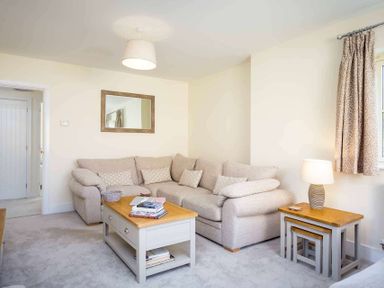 A living room with a beige sofa a coffee table with books a side table with a lamp and a window with curtains at The Bybrook in Bourton-on-the-Water