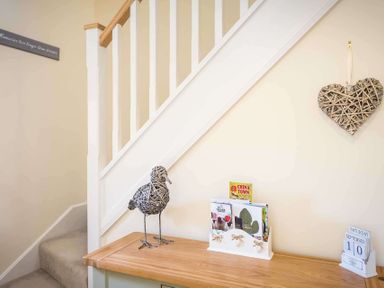 A staircase with white banisters above a wooden table holding a wicker bird, magazine holder, calendar and a wicker heart on the wall at The Bybrook in Bourton-on-the-Water