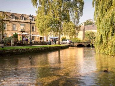 A river with trees and ducks next to a stone building and a stone bridge at The Bybrook in Bourton-on-the-Water