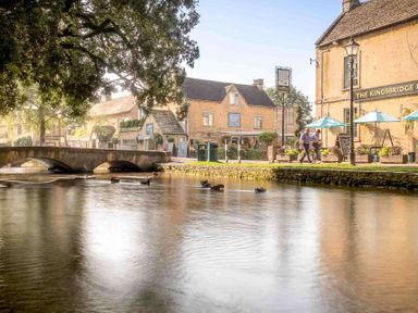 A river with ducks and a stone bridge next to buildings and people walking at The Bybrook in Bourton-on-the-Water