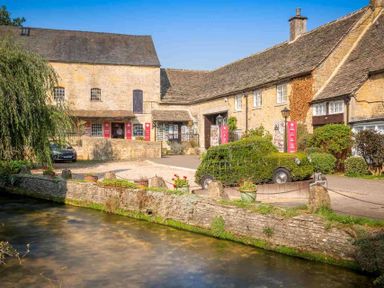 Stone buildings by a river with a green topiary shaped like a car at The Bybrook in Bourton-on-the-Water