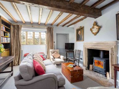 A living room with a sofa and armchair facing a wood burning stove with a wooden coffee table and exposed wooden ceiling beams at Shenley Broadwell