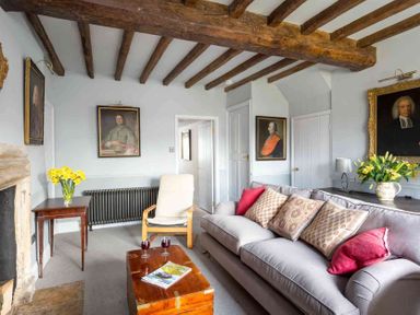 A living room with a gray sofa with cushions a wooden chair a wooden coffee table with two glasses and a magazine a side table with yellow flowers and framed portraits on the walls at Shenley in Broadwell