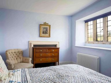 A bedroom with a quilted bedspread a wooden chest of drawers an armchair a framed picture and a window with wooden panes at Shenley in Broadwell