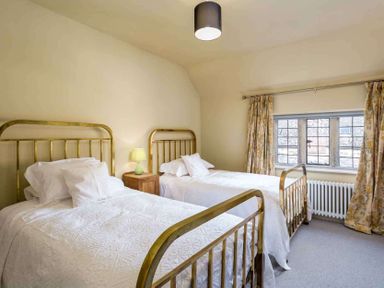 A bedroom with two single beds with brass frames a wooden nightstand with a lamp and a window with floral curtains at Shenley in Broadwell
