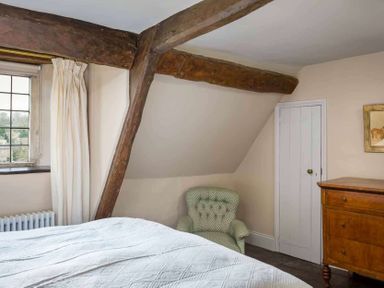A bedroom with wooden beams a green armchair a wooden dresser a white bedspread and a window with curtains at Shenley in Broadwell