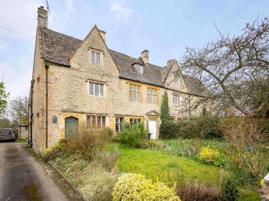 A stone house with a sloped roof surrounded by bushes and trees with a grass lawn at Shenley Broadwell