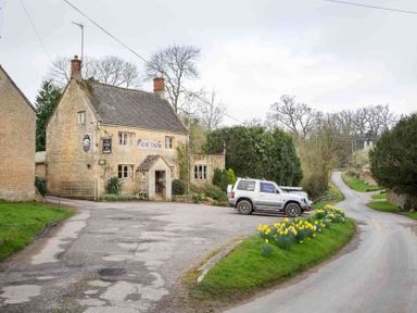 A stone building labeled Fox Inn next to a small parking area with a car and a road lined with flowers at Shenley in Broadwell