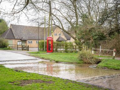 A rural scene with a shallow water crossing a road a red telephone box and a building surrounded by trees at Shenley Broadwell