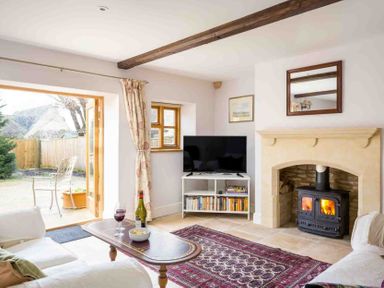 A living room with a wood burning stove TV on a white stand and an open door to a garden at Oyster Barn in Naunton