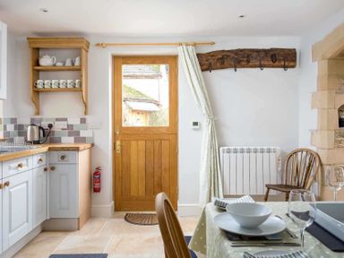 A kitchen with wooden door and shelf with mugs next to a dining table set with plates glasses and cutlery at Oyster Barn in Naunton