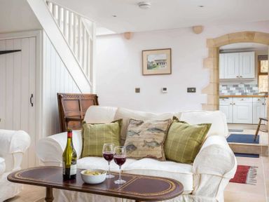 A living room with a white sofa and green pillows a wooden table with wine and snacks and a view into a kitchen at Oyster Barn in Naunton